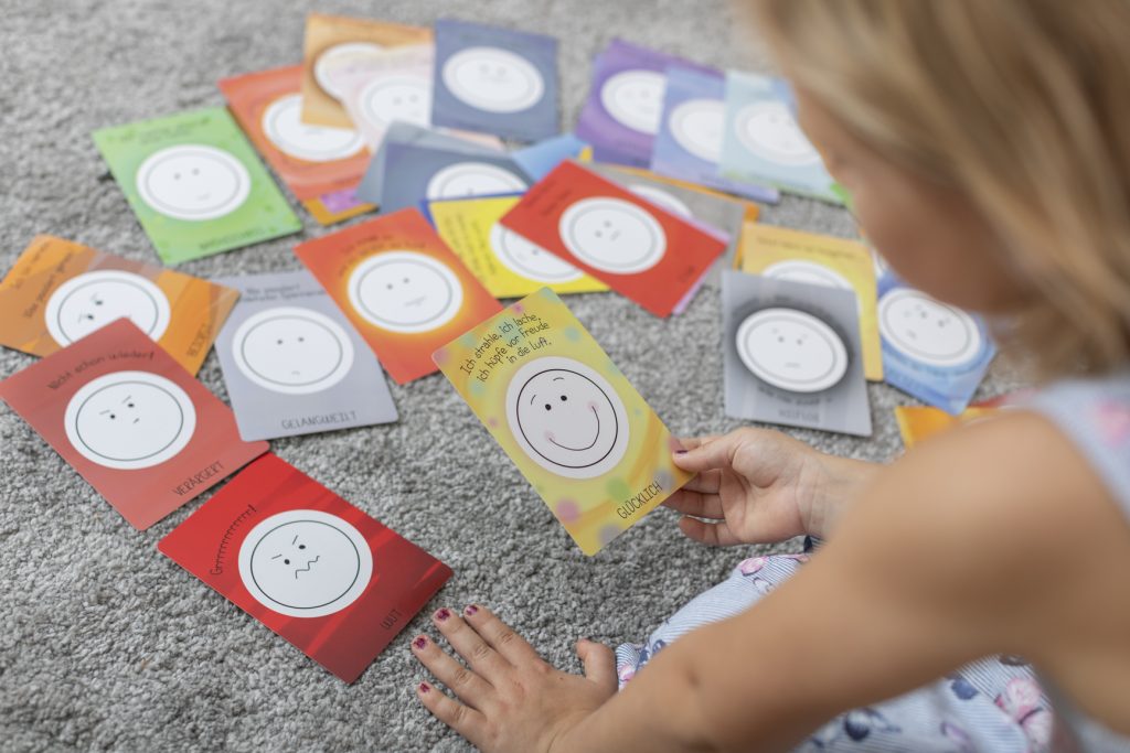 Ein Mädchen betrachtet verschiedene Emotionskarten, die auf einem Teppich verteilt liegen.