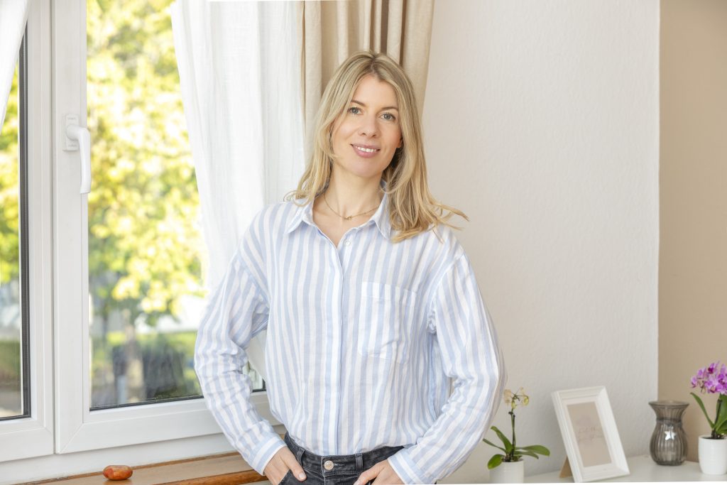 Die Therapeutin Judith Mack in ihren Behandlungsräumen vor dem Fenster stehend, die Hände locker in die Hosentaschen gesteckt. Auf dem Sideboards rechts sind ein Bilderrahmen, zwei Orchideen und eine Vase erkennbar.